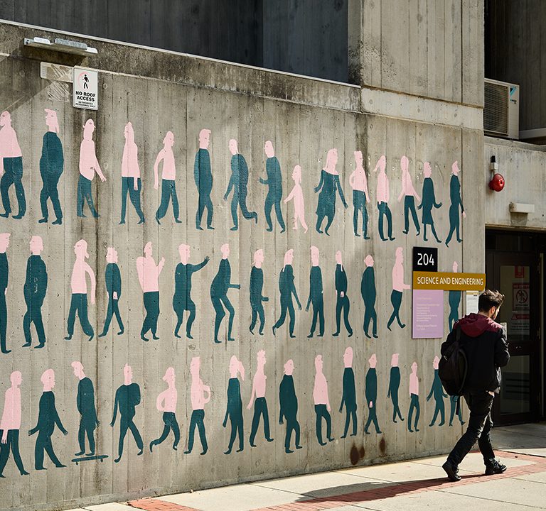 A student walks past a cement wall with an artwork that shows people painted in pink and green.