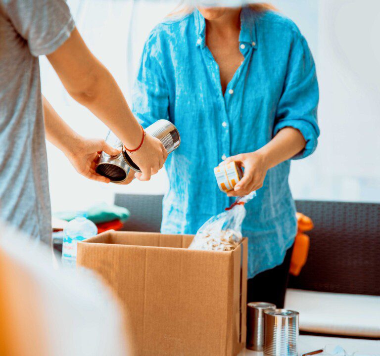 A person hands two unlabelled cans of food to a volunteer in a bright blue shirt. The photo shows them from the neck down.
